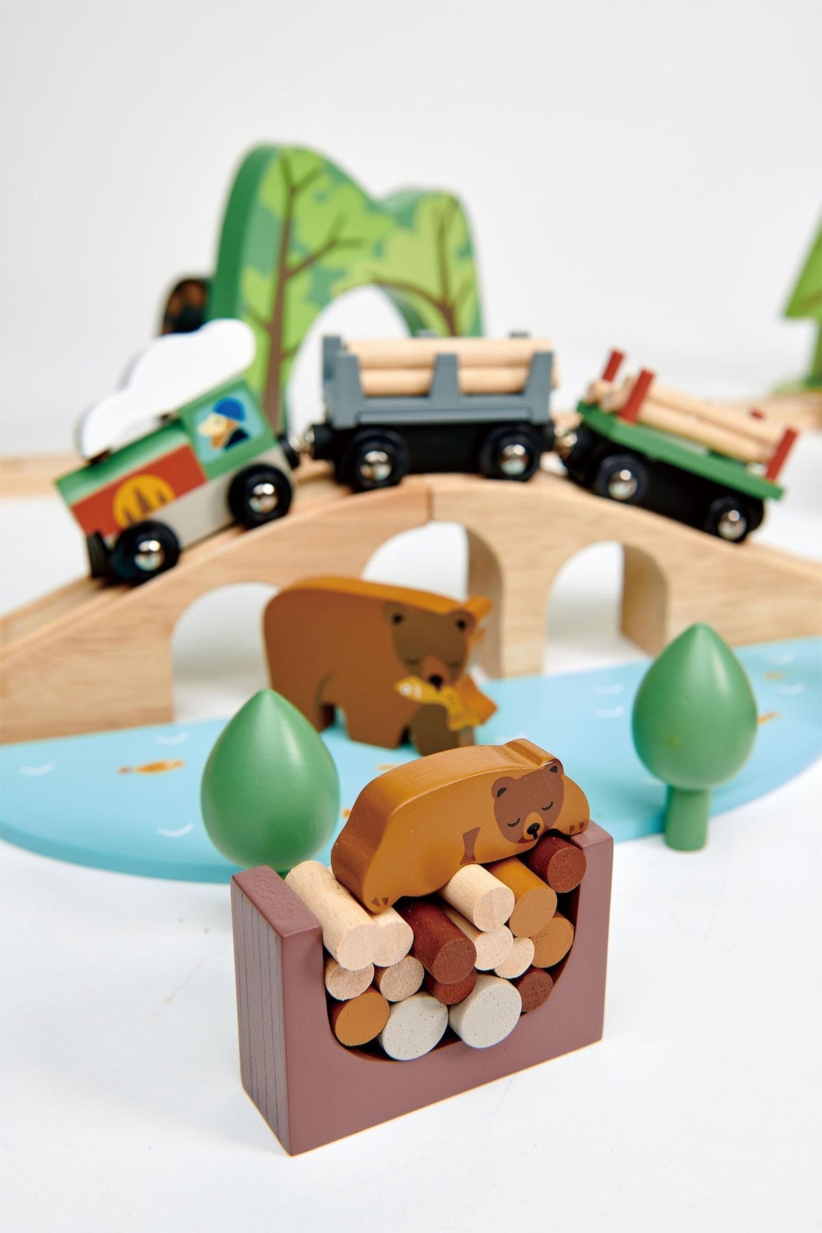 A colorful wooden train set features two playful bear figures and a log pile, set against a backdrop of stylized trees and a bridge, encouraging imaginative play.