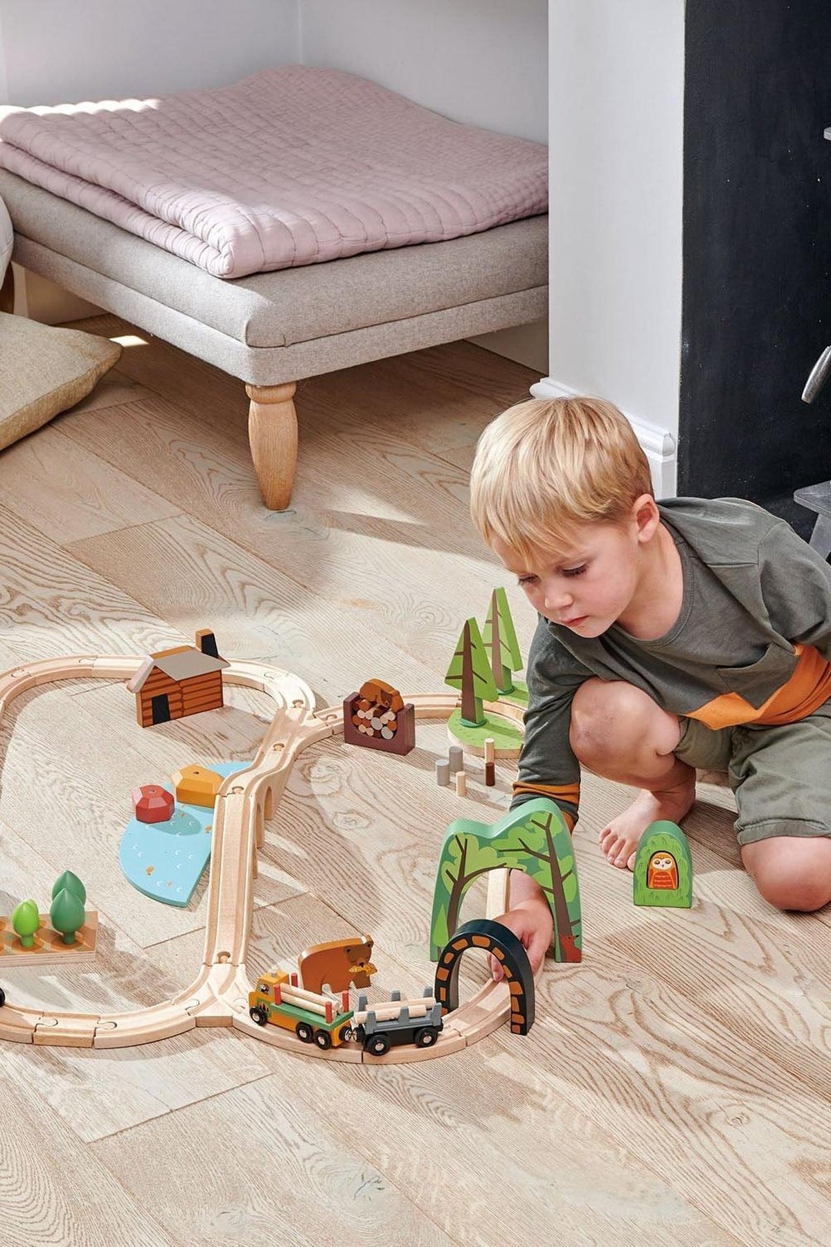 A young boy plays with the Tender Leaf Wooden Toys Wild Pines Train Set on a light wooden floor, featuring colorful logs, trees, and a tunnel, promoting imaginative storytelling.