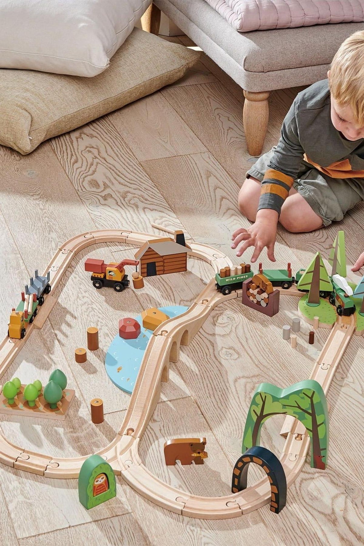 A young child plays with a colorful wooden train set on a light wooden floor, featuring trees, a cabin, and vehicles, promoting imaginative storytelling and sustainable play.