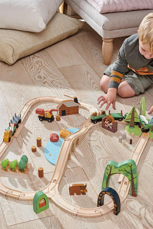 A young child plays with a colorful wooden train set on a light wooden floor, featuring trees, a cabin, and vehicles, promoting imaginative storytelling and sustainable play.