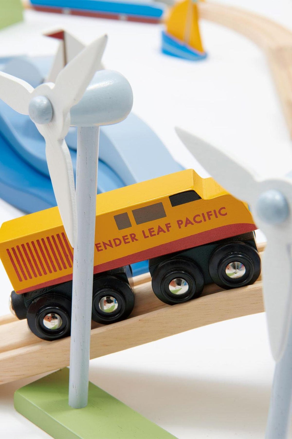 A colorful wooden train with the label Tender Leaf Pacific travels along a track surrounded by whimsical wind turbines and vibrant landscapes, showcasing eco-friendly design and imaginative play.