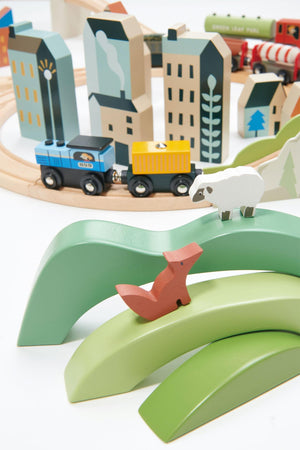 Colorful wooden animals and hills are positioned in front of a train set with vibrant cars, surrounded by stylized buildings and trees, creating an engaging and imaginative play environment.
