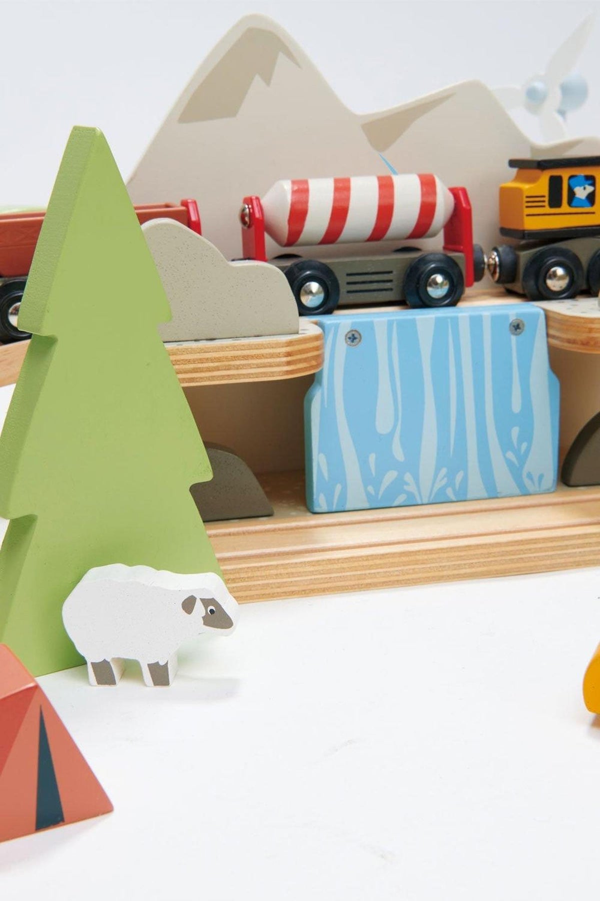 A colorful wooden train set features various vehicles, a blue water feature, and playful scenery including a green tree, white sheep, and geometric tents, perfect for imaginative play.