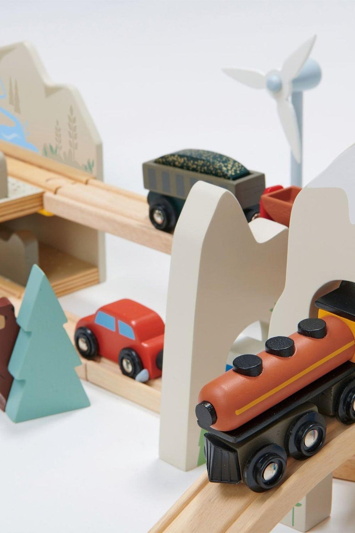 Colorful wooden vehicles and scenic elements are arranged on a train track, featuring a black and orange train, a red car, and green trees amidst a playful mountain landscape.