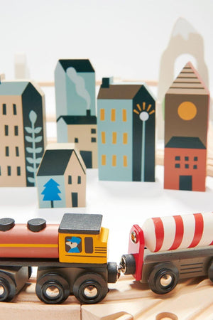 A colorful wooden train set features a vibrant engine and tanker, set against a backdrop of stylized wooden buildings in various shapes and colors, promoting imaginative play.