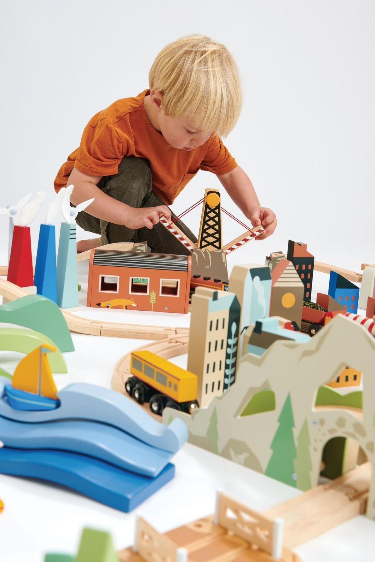 A young child plays with a colorful wooden train set, featuring various buildings, trees, and a bridge, arranged on a light surface.