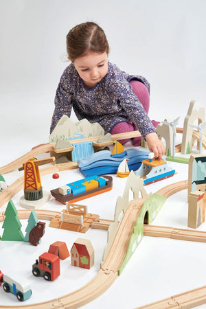 A child engages with a colorful wooden train set, featuring a river, mountains, and various vehicles. The eco-friendly materials promote imaginative play, perfect for young children.