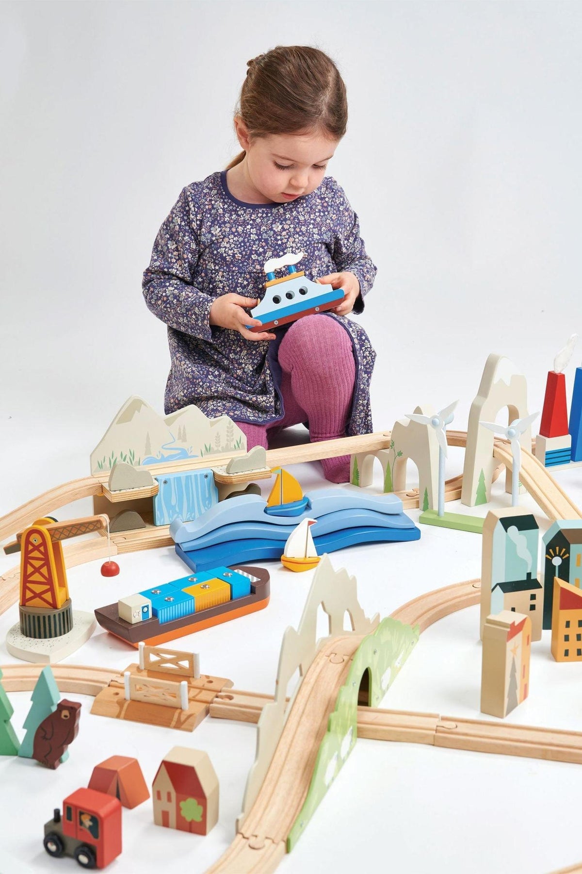A young girl sits on the floor, playing with a colorful wooden train set featuring mountains, a river, and various buildings.