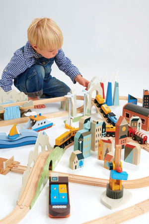 A young child plays with a colorful wooden train set featuring various buildings, vehicles, and landscapes. The eco-friendly toys encourage imaginative play and are made of sustainable materials.