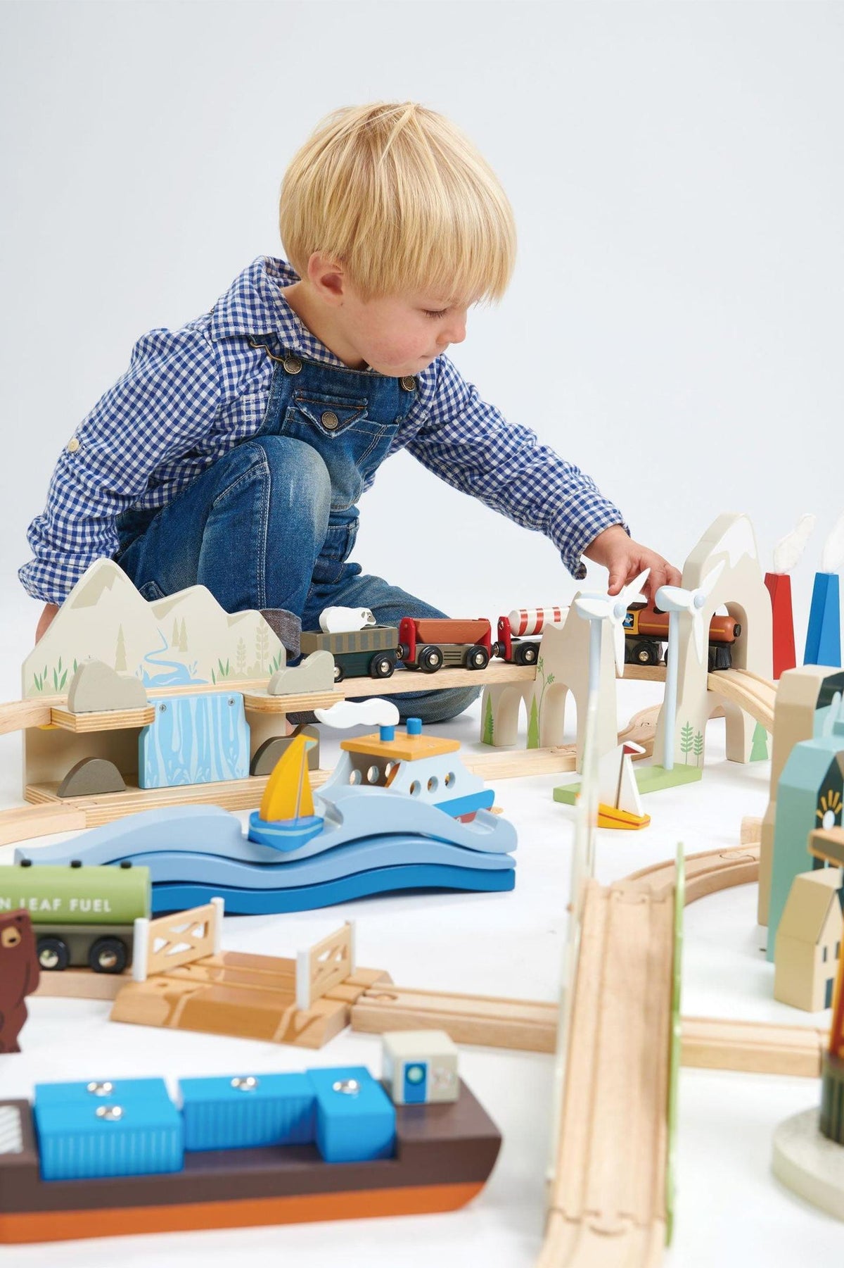 A young child plays with a colorful wooden train set surrounded by various landscape features, including mountains, water, and buildings.
