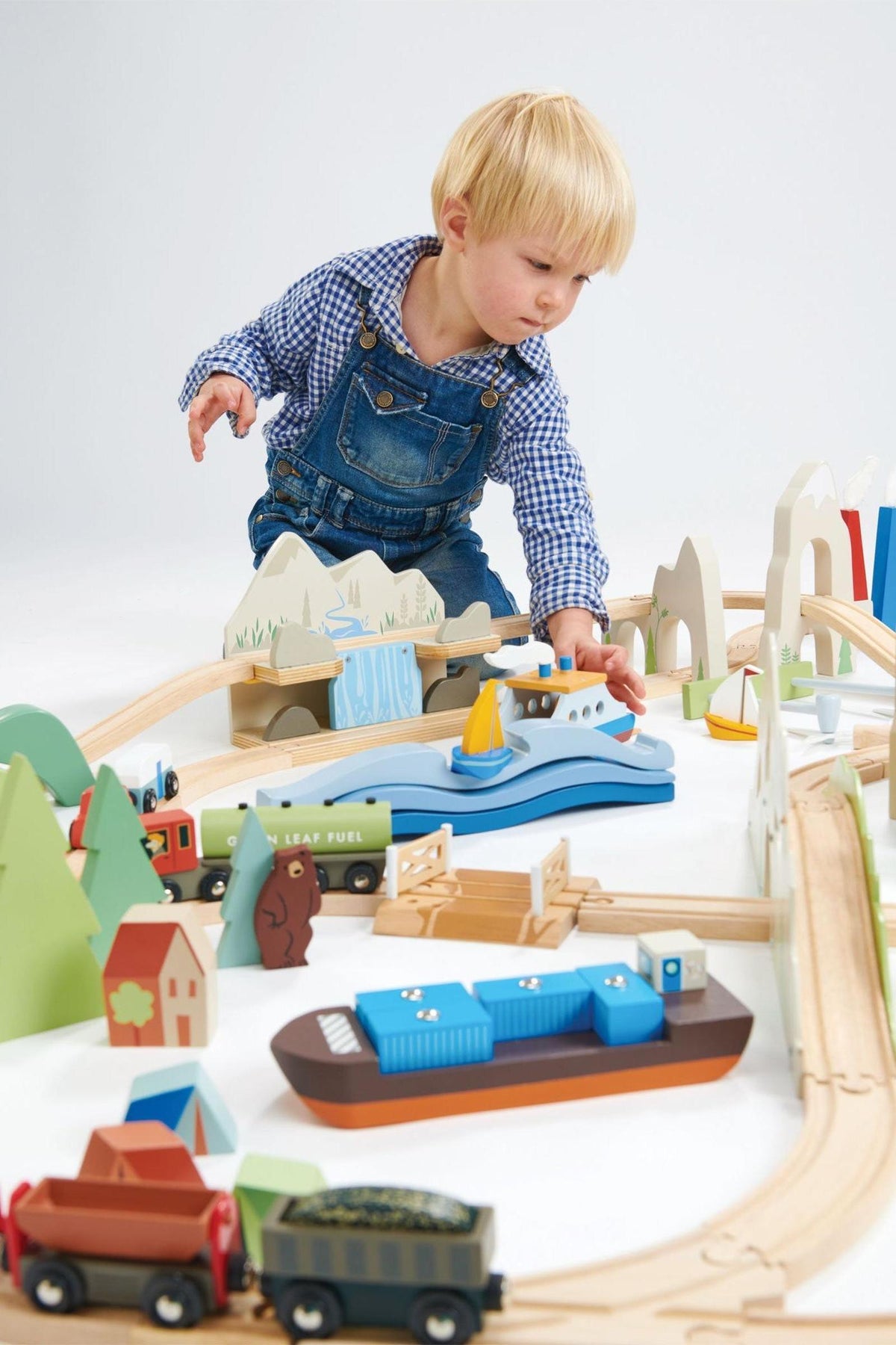A young child plays with a colorful wooden train set, featuring a blue boat and various vehicles, surrounded by trees and mountains, on a light-colored surface.