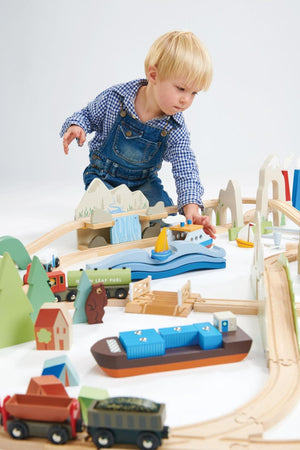 A young child plays with a colorful wooden train set, featuring a blue boat and various vehicles, surrounded by trees and mountains, on a light-colored surface.