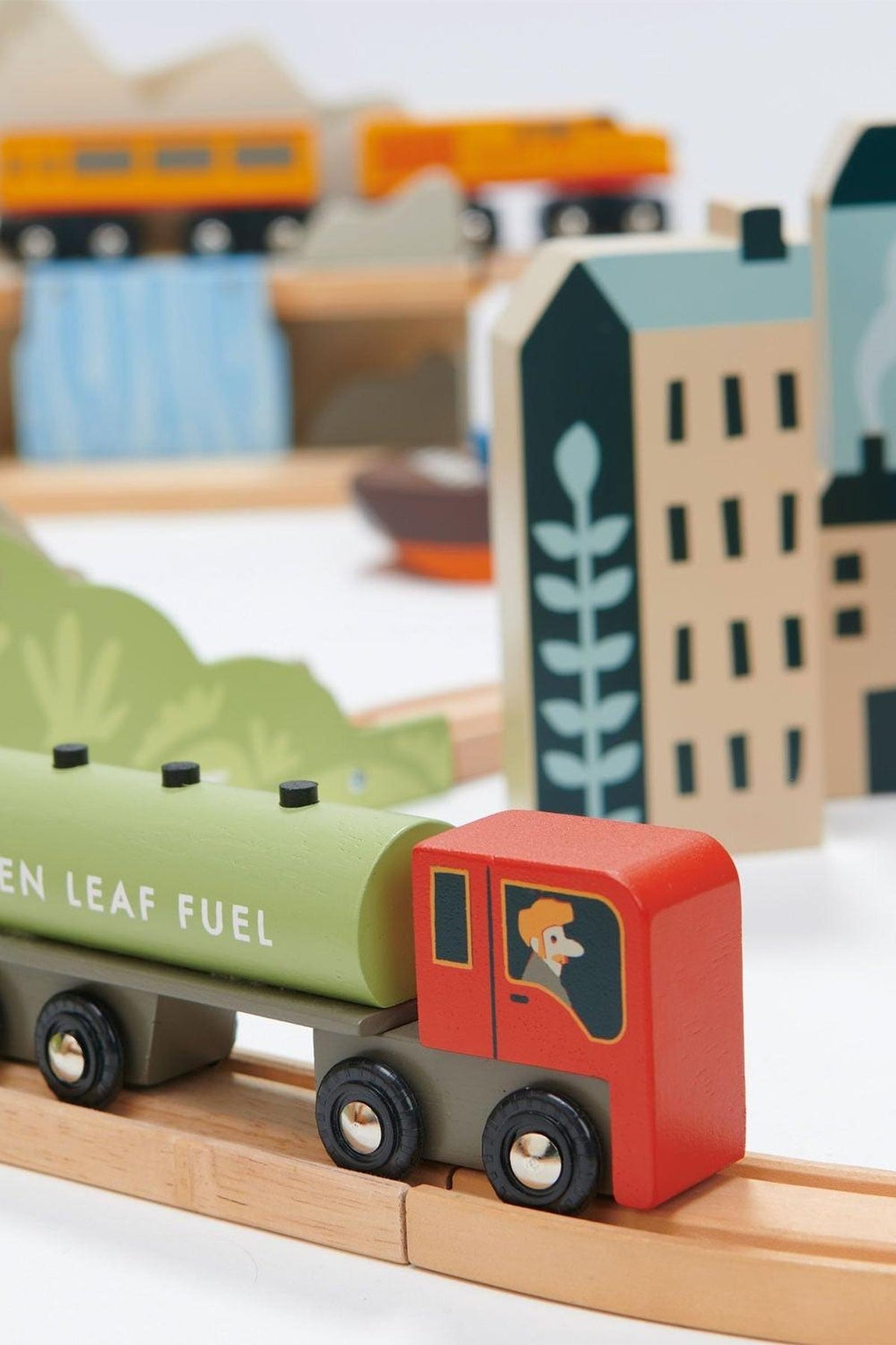 A colorful wooden train set features a green fuel tanker with a smiling engineer, surrounded by stylized buildings and landscapes, designed for imaginative play with sustainable materials.