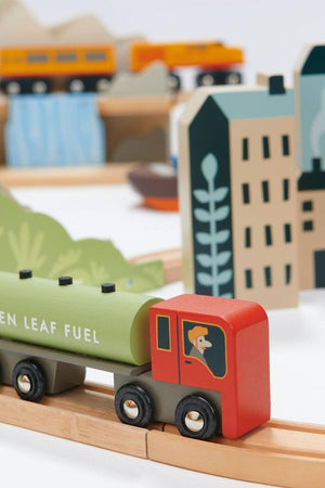 A colorful wooden train set features a green fuel tanker with a smiling engineer, surrounded by stylized buildings and landscapes, designed for imaginative play with sustainable materials.