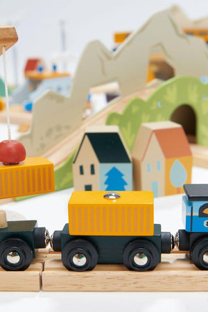 A colorful wooden train set featuring a black locomotive and yellow cargo containers, set against a backdrop of stylized wooden houses and green hills, promoting imaginative play for young children.