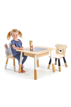 A young girl sits at a light wooden table with a removable chalkboard panel, flanked by two animal-themed chairs featuring bear and bunny faces, emphasizing its playful design for toddlers.
