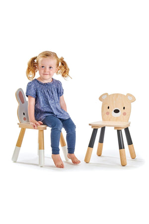 A young girl sits on a wooden chair with a bunny face back, beside another chair featuring a bear face. Both chairs are sustainably made, with a natural wood finish.