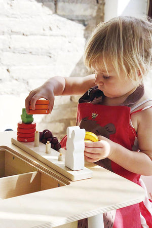 A toddler with short blonde hair plays with colorful stacking toys on a wooden activity table. The child is focused, wearing a red apron,.