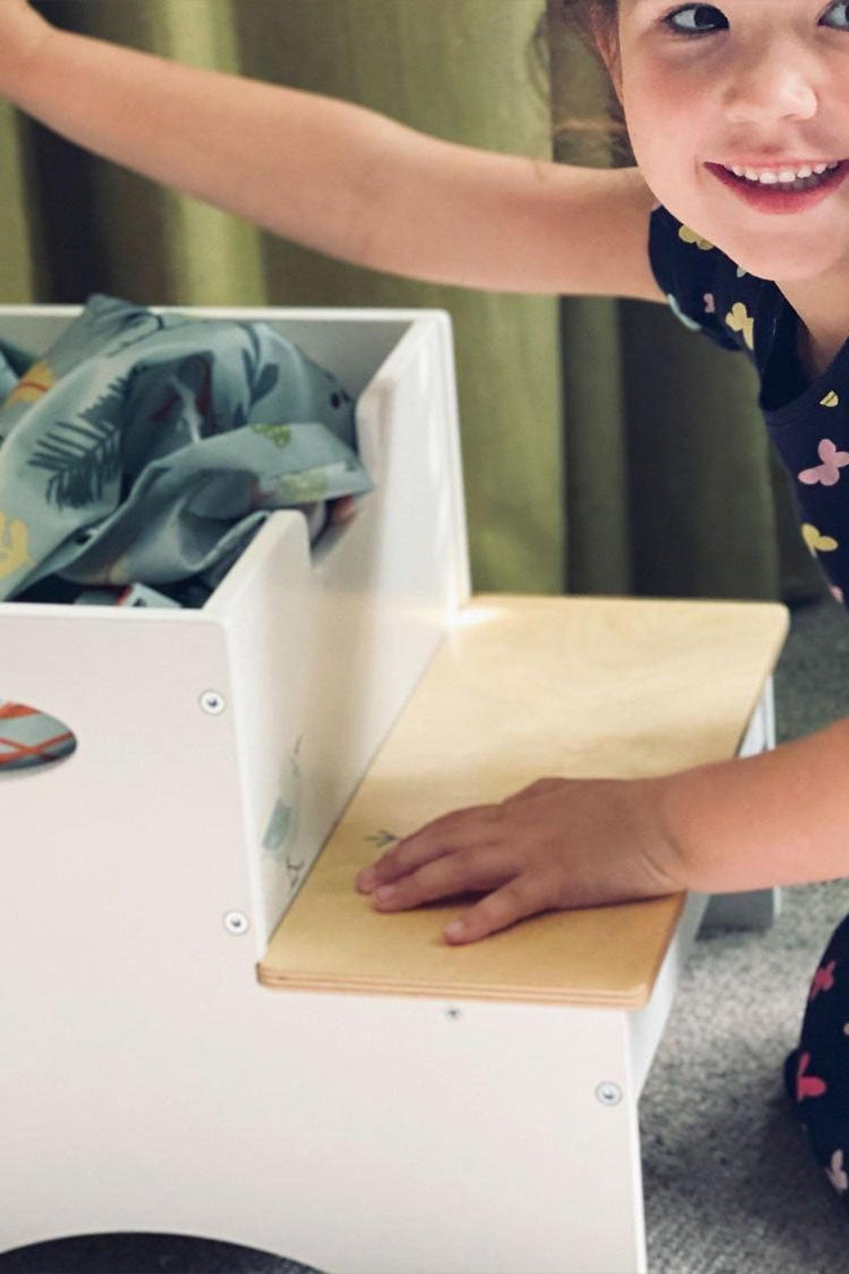 A smiling child sits near a white wooden stool with a natural wood top, reaching inside for toys. The stool features easy-grip handles and a hidden storage compartment.