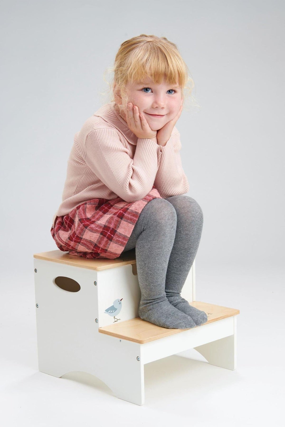 A young girl sits on a two-step stool with a natural wood top and white body, wearing a pink sweater and plaid skirt.