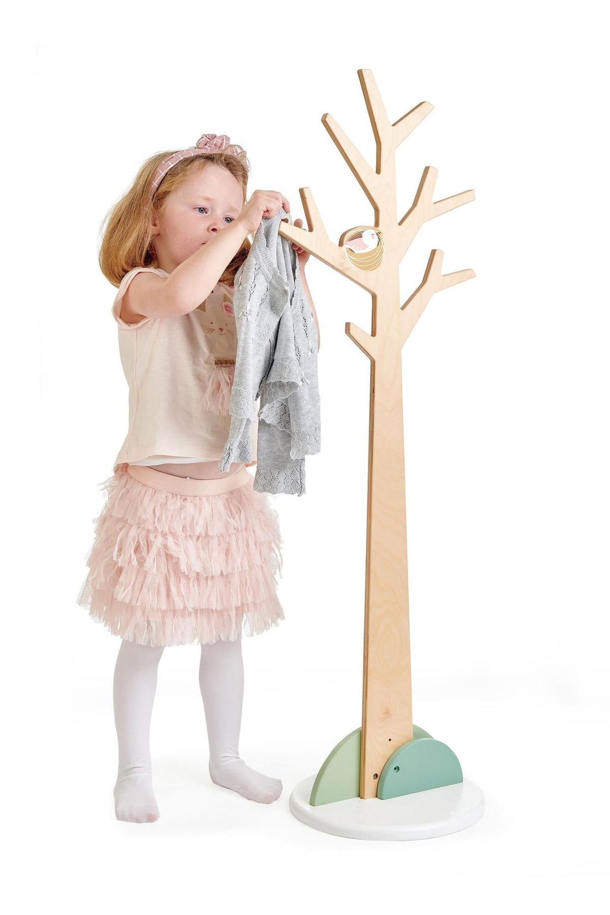 A young girl in a pink skirt hangs a gray sweater on a natural wood coat stand designed for children.