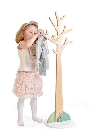 A young girl in a pink skirt hangs a gray sweater on a natural wood coat stand designed for children.