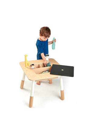 A toddler plays with toys on a wooden table with a removable chalkboard panel, revealing storage compartments.