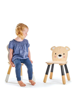 A young girl sits on a wooden chair, facing a bear-shaped chair with a friendly face, showcasing sustainable design.
