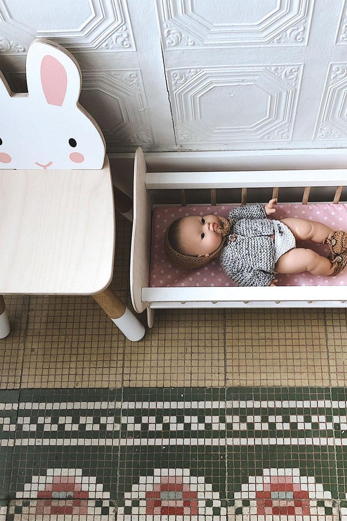 A wooden toddler chair with a rabbit design sits beside a doll in a miniature bed on a patterned floor.