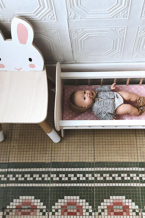 A wooden toddler chair with a rabbit design sits beside a doll in a miniature bed on a patterned floor.