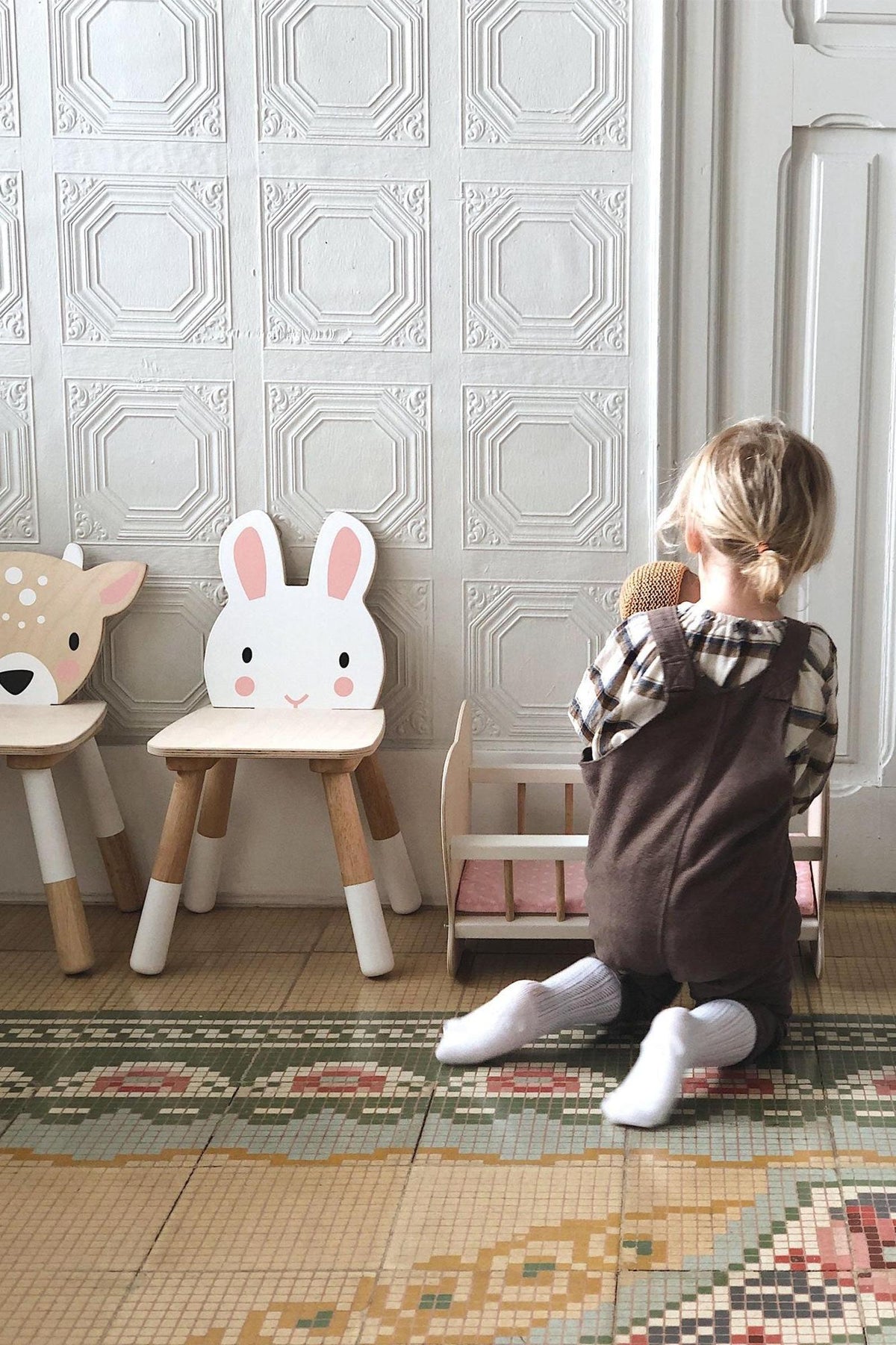 A child kneels on a patterned floor, playing with a soft toy beside two wooden animal chairs, including a rabbit.