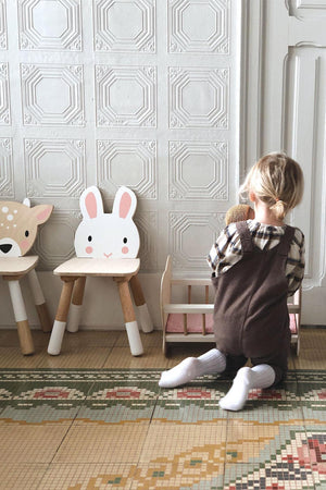 A child kneels on a patterned floor, playing with a soft toy beside two wooden animal chairs, including a rabbit.
