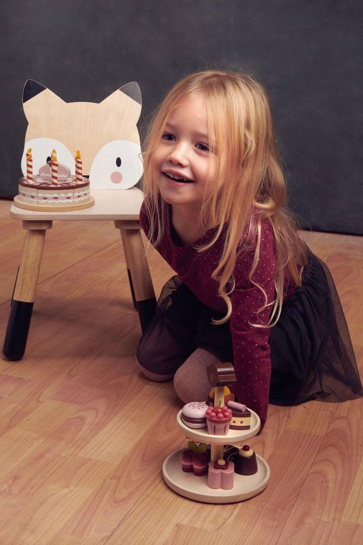A smiling child with long hair kneels beside a wooden fox chair, playing with a colorful wooden dessert display.
