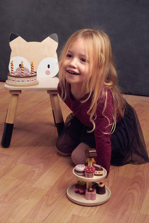 A smiling child with long hair kneels beside a wooden fox chair, playing with a colorful wooden dessert display.