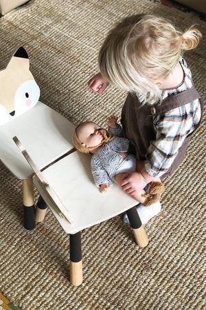 A toddler sits beside a fox-shaped wooden chair, gently holding a doll dressed in a knitted outfit on the floor.