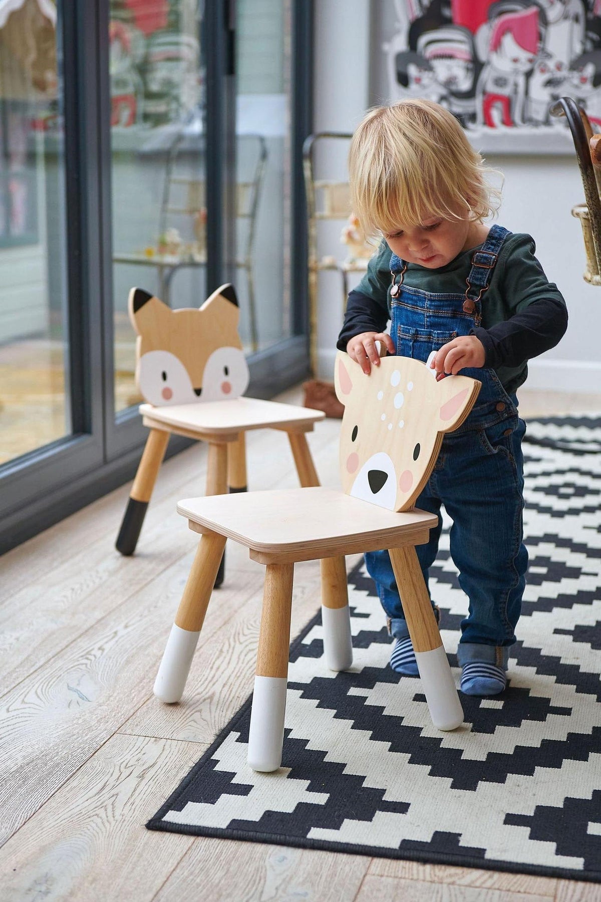 A toddler in overalls interacts with a wooden deer chair, featuring a playful design. Two similar animal-themed chairs are visible on a patterned rug in a bright indoor setting.
