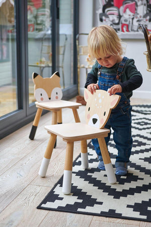 A toddler in overalls interacts with a wooden deer chair, featuring a playful design. Two similar animal-themed chairs are visible on a patterned rug in a bright indoor setting.