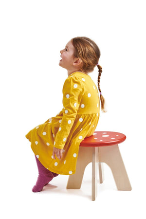 A child in a yellow polka dot dress sits on a wooden toadstool stool with a red top, smiling joyfully.