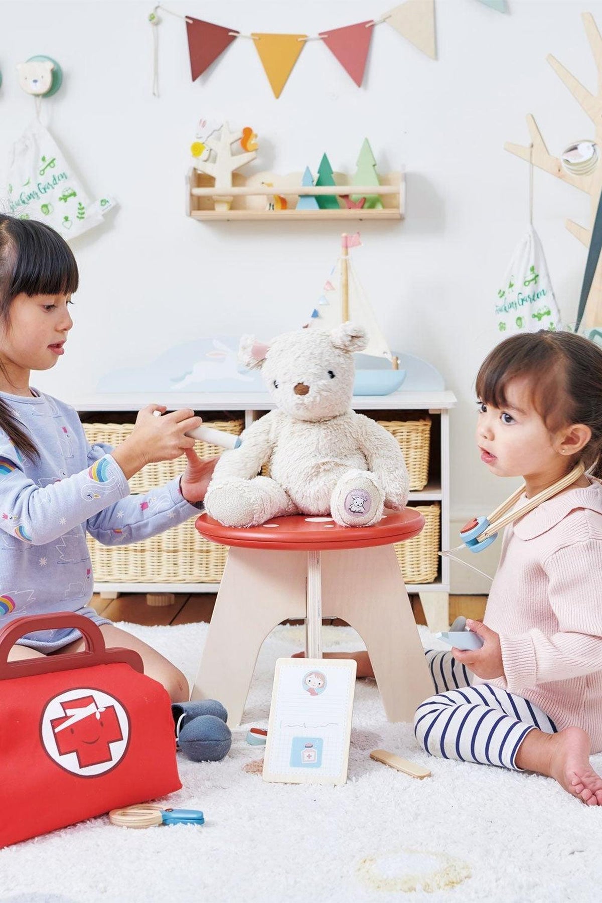 Two children play with a stuffed bear on a red toadstool stool in a bright, playful nursery filled with toys.