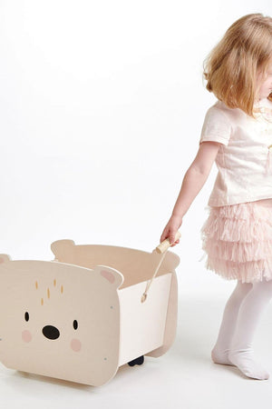 A young child pulls a light-colored wooden bear cart with a friendly face, designed for carrying toys and books.