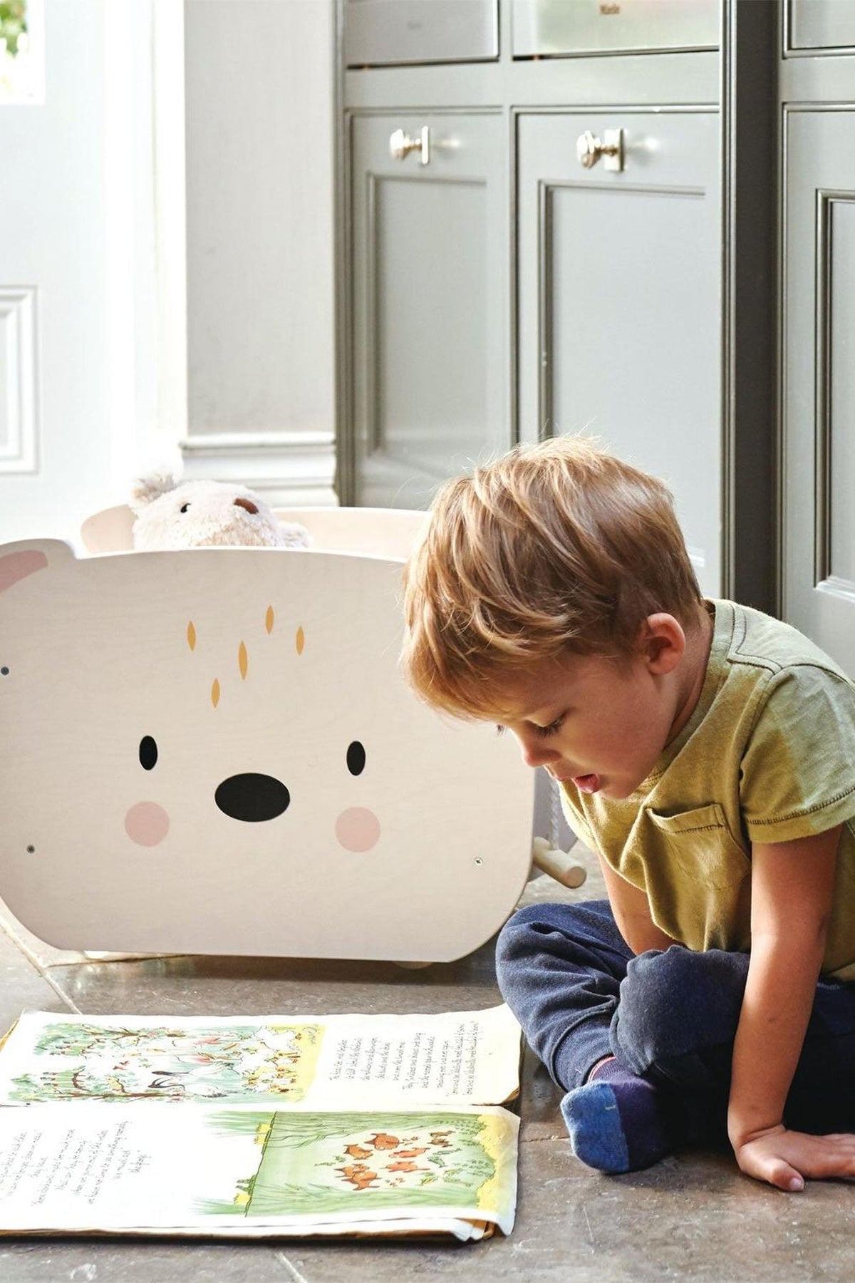 A young child sits on the floor, reading a book in front of a bear-shaped toy cart made of wood.