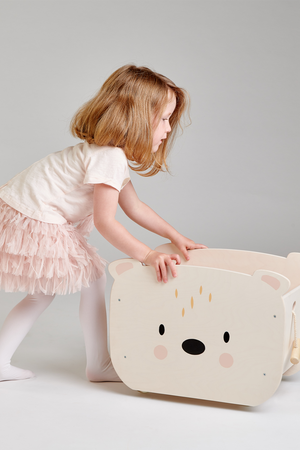 A young child with blonde hair in a light shirt and pink tutu is interacting with a wooden bear-shaped cart.