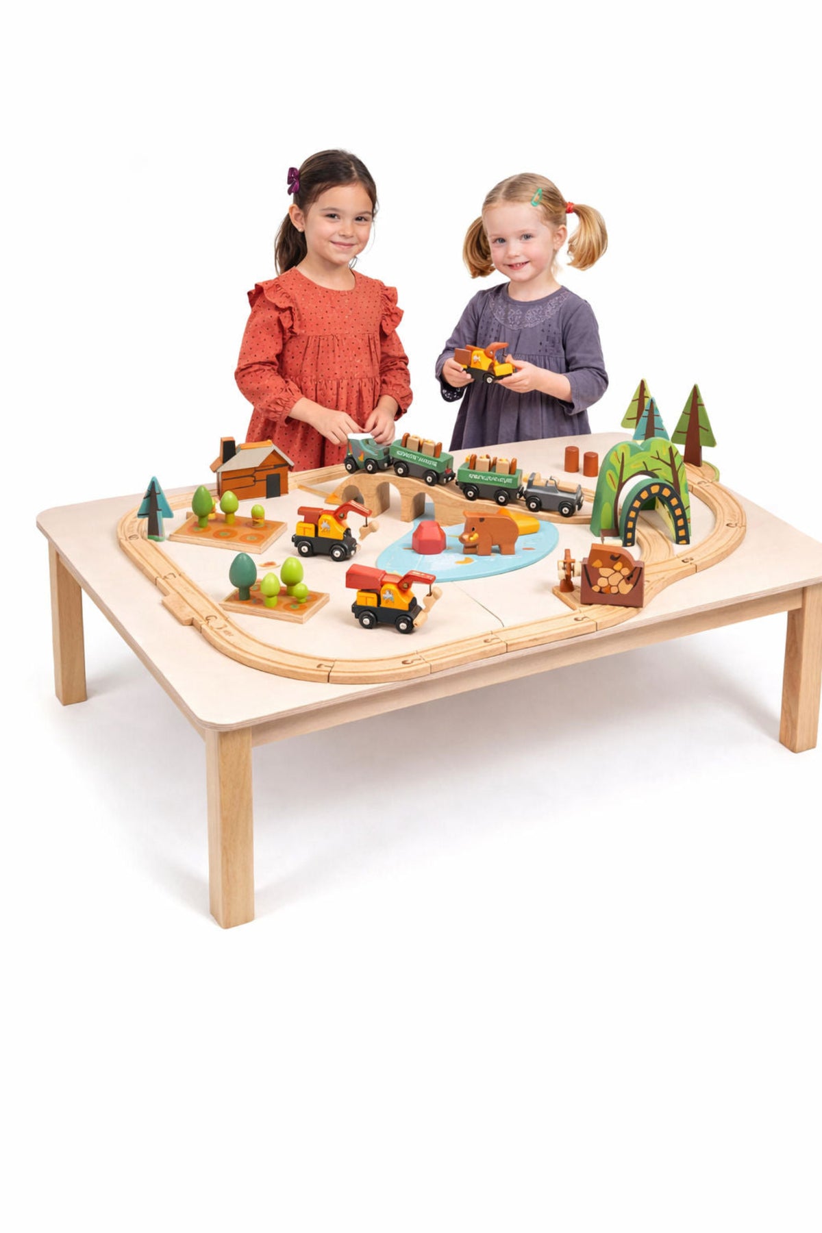 Two children play together at a light wooden table, surrounded by vibrant toy trains, trees, and scenic elements, engaging in creative play with a colorful train set.