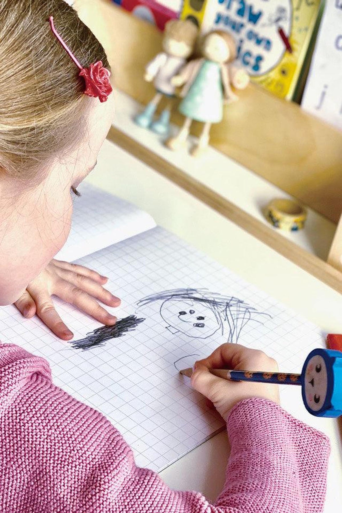A young child with a pink sweater draws in a notebook, surrounded by colorful toys and books, showcasing creativity and engagement in a playful, bright environment.