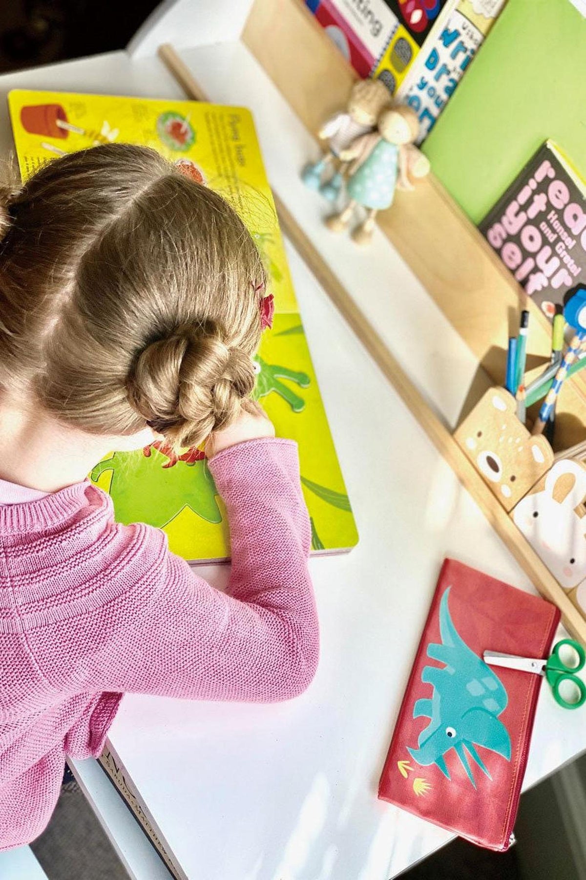 A young child with a braided hairstyle is focused on a colorful activity book on a white desk, surrounded by playful stationery and decorative items in a bright, cheerful setting.
