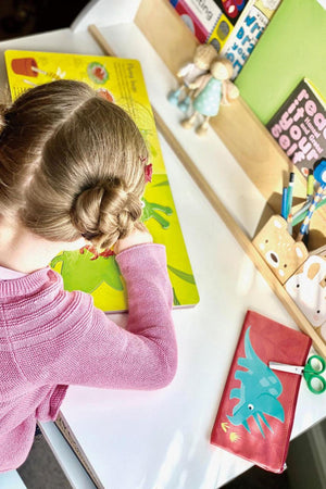 A young child with a braided hairstyle is focused on a colorful activity book on a white desk, surrounded by playful stationery and decorative items in a bright, cheerful setting.