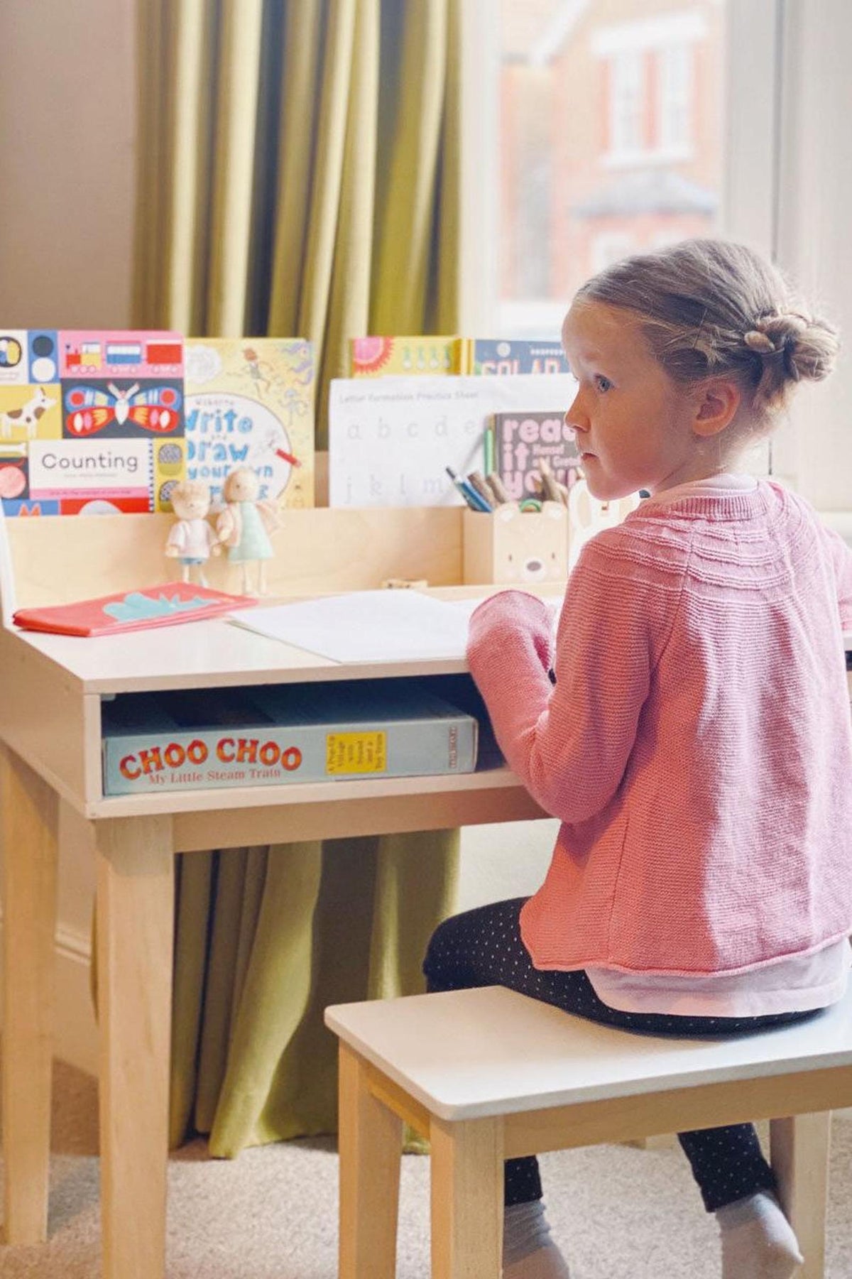 A young child sits at a wooden desk, wearing a pink sweater, with colorful educational materials visible in the background.