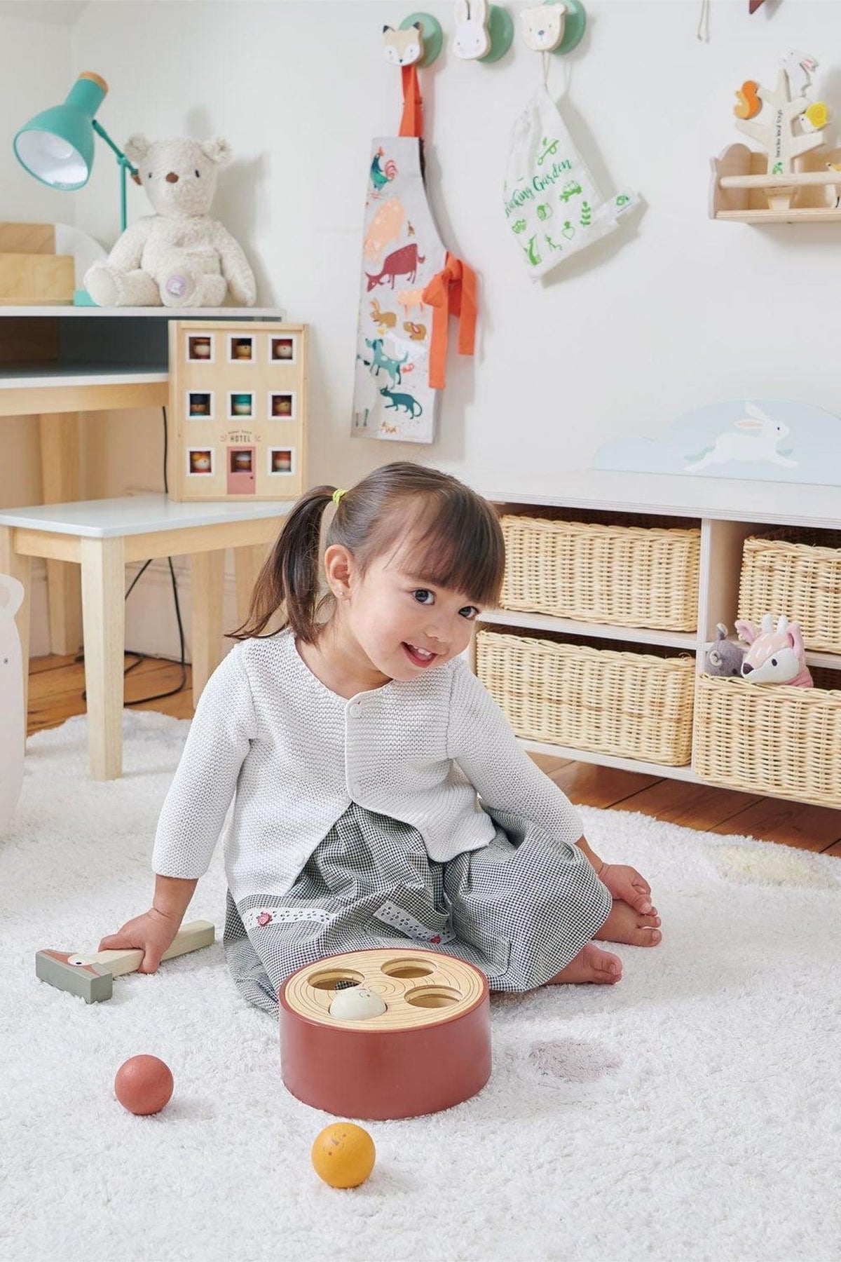 A young child sits on a soft rug in a bright, playful room, engaging with a wooden toy while surrounded by woven storage baskets and colorful wall decor.