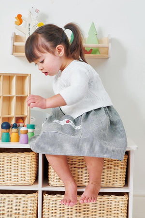 A young child in a white sweater and checkered skirt plays with colorful wooden figures on a storage unit, surrounded.