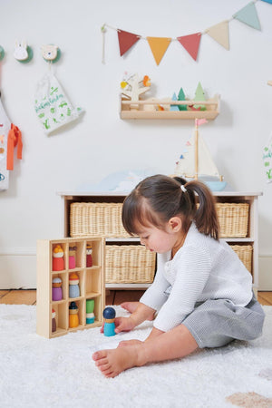 A young child plays on a soft rug, interacting with colorful wooden figures displayed in a natural wood storage box.